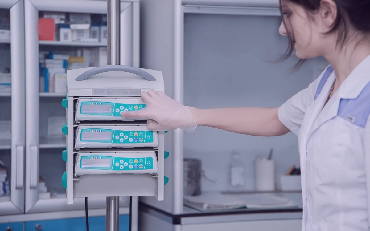 Clinician adjusting infusion pump controls beside medical supply cabinets.